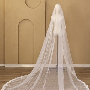 Puede incluir: Un velo de novia largo y blanco con delicados bordes de encaje. El velo se exhibe en un maniqu&iacute;, mostrando su dise&ntilde;o fluido y sus intrincados detalles. El ribete de encaje a&ntilde;ade un toque de elegancia al accesorio de boda.