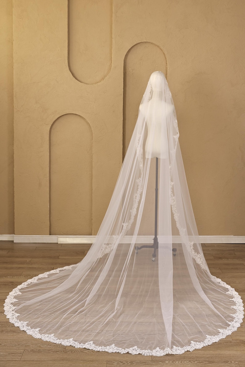 Puede incluir: Un velo de novia largo y blanco con delicados bordes de encaje. El velo se exhibe en un maniqu&iacute;, mostrando su dise&ntilde;o fluido y sus intrincados detalles. El ribete de encaje a&ntilde;ade un toque de elegancia al accesorio de boda.