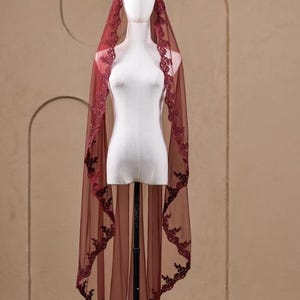 K&ouml;nnte beinhalten: Ein bordeauxfarbener Brautschleier mit aufwendiger floraler Spitzenborte. Der transparente Stoff drapiert sich &uuml;ber einer Schaufensterpuppe und zeigt das filigrane Design. Der Schleier hat eine tiefrote Farbe, wobei die Spitze einen kontrastierenden dunkleren Farbton hinzuf&uuml;gt.
