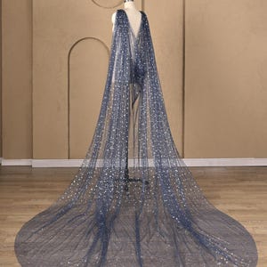 May include: A floor-length, sheer navy blue cape made of tulle fabric, featuring a V-neckline and a scattered pattern of silver glitter stars. The cape drapes from the shoulders, creating a long, flowing train that pools on the floor.