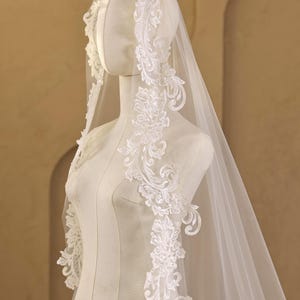 Pode incluir: Um véu de noiva branco com detalhes intrincados de renda floral nas bordas. O véu é drapeado sobre um manequim, mostrando seu design delicado e tecido transparente. A renda apresenta padrões florais detalhados.