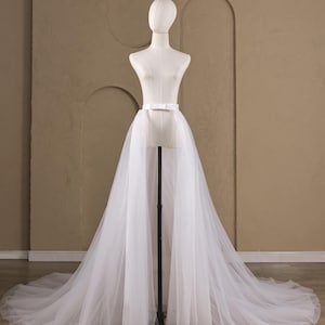 May include: A white tulle overskirt featuring a long, flowing train and a structured waistband with a small bow detail at the center front. The sheer, layered fabric creates a voluminous silhouette that extends to the floor, displayed on a mannequin.