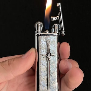 以下が含まれることがあります： 前面に剣のデザインが施された銀色のライター。ライターに火が灯り、上部から炎が出ている。