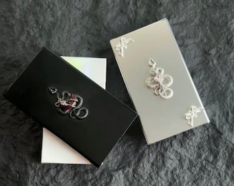 Könnte beinhalten: Zwei kleine rechteckige Schachteln mit einem Schlangendesign. Eine Schachtel ist schwarz mit einem roten Stein und die andere ist silber mit einem rosa Stein.