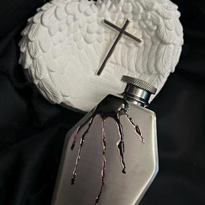 May include: A silver metal flask shaped like a coffin with a black liquid dripping down the side. The flask is in front of a white plaster sculpture of angel wings with a cross.