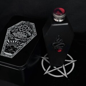 Puede incluir: Frasco negro en forma de ataúd con una gema roja y un diseño de serpiente. El frasco viene en una lata negra con un diseño de tablero Ouija plateado. El texto en el tablero Ouija dice "Hello, Goodbye, ABCDEFGHIJKLMNOPQRSTUVWXYZ, Yes, No".