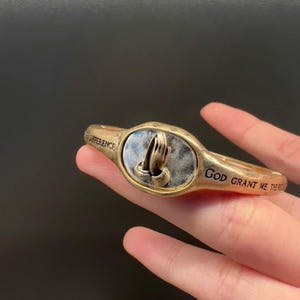 Könnte beinhalten: Goldfarbenes Armband mit einem silbernen ovalen Anhänger mit einem Motiv von betenden Händen. Das Armband trägt die Inschrift "God Grant Me The Strength To Make A Difference" auf dem Band.