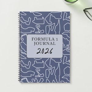 May include: A spiral-bound notebook with a blue cover featuring white outlines of race tracks. The notebook has a rectangular label that reads "FORMULA 1 JOURNAL 2026". The notebook is on a white surface.