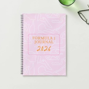 Op de afbeelding: Een roze spiraalgebonden notitieboek met een lichtroze achtergrond met witte contouren van racecircuits. Het notitieboek heeft de tekst "FORMULA 1 JOURNAL 2026" in goudkleur. Een bril en een glas water staan op de achtergrond.