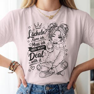 May include: A light pink t-shirt featuring a black line art graphic of a person with double buns, wearing a sweater and ripped jeans, giving the middle finger. The text reads: L&auml;cheln? Kann ich. Muss ich ABER NICHT! Deal with it.