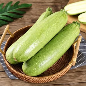 May include: Three fresh, light green courgettes with smooth, speckled skin are arranged inside a round, woven rattan basket featuring two small wooden handles. The vegetables are cylindrical in shape with tapered ends and a firm, fresh appearance.