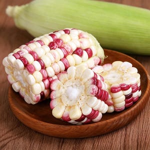 Può includere: Un pezzo di pannocchia di mais bicolore con un mix di chicchi bianchi e rosso scuro, accompagnato da due sezioni trasversali dello stesso mais, il tutto su un piccolo piatto di legno con una pannocchia intera e sbucciata sullo sfondo.