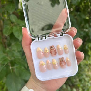 May include: A set of almond-shaped press-on nails in a clear plastic case. The nails have a light pink base with yellow plaid tips and brown bear accents. The case is held in a hand against a blurred green background.