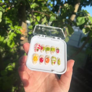 May include: A set of press-on nails with fruit-themed designs. The nails feature illustrations of strawberries, apples, oranges, pears, and a smiling sun. The nails are in a white plastic case.