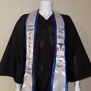 May include: Black graduation gown with white and blue accents. The white sash displays the text "Galen College of Nursing" and "First Gen" on one side, and the name "Kaitlyn Rieu", "BSN", and "RN Class of 2023" on the other. A caduceus symbol is also visible.