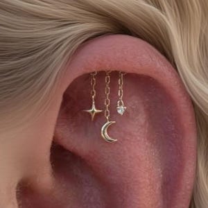 May include: Close-up of an ear with three gold chain earrings. One earring has a star, another a crescent moon, and the third a small diamond-shaped charm. The earrings are attached to the ear with small posts.