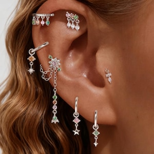 May include: A collection of silver earrings and ear cuffs with colorful gemstones. The earrings include hoops with star charms and dangling gems, and studs with floral and geometric designs. The ear cuffs feature dangling gems.