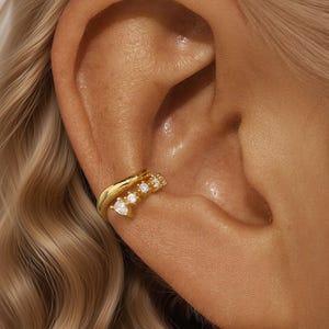 May include: A gold ear cuff with a curved design. The cuff features several clear, sparkling stones, including a heart-shaped stone. The jewelry is worn on the ear and is set against a background of blonde hair.