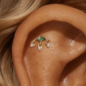 Puede incluir: Un piercing de oro con una piedra cuadrada verde y tres gemas colgantes en forma de lágrima con piedras transparentes. La joya está colocada sobre el fondo de una oreja.