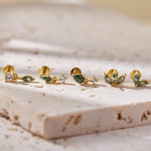May include: Several gold stud earrings are displayed. Some have clear, round gemstones and green, teardrop-shaped stones. Others feature a floral design with green and white accents. The earrings are presented on a light-coloured, textured surface. The earrings are made of gold.