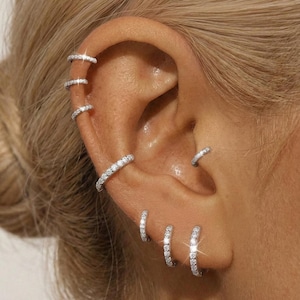 May include: Close-up of an ear adorned with multiple silver hoop earrings. The earrings are encrusted with small, sparkling stones, creating a dazzling effect. The ear has multiple piercings, showcasing the variety of earring styles. The overall aesthetic is elegant and glamorous.