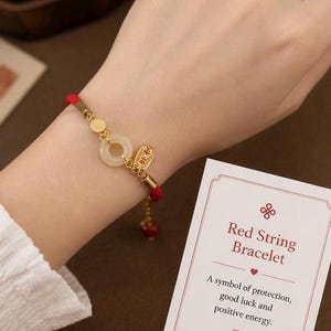 May include: Red string bracelet with gold accents and a jade pendant. The bracelet is on a wrist, next to a card that reads "Red String Bracelet" and describes it as a symbol of protection, good luck, and positive energy.