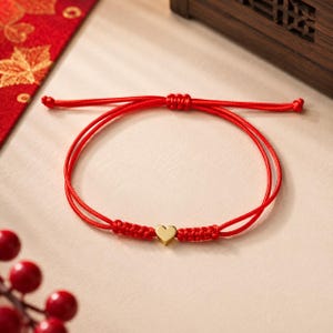 May include: A red cord bracelet featuring a small, polished gold-tone heart charm at the center. The bracelet is constructed with thin, vibrant red string and includes an adjustable sliding knot mechanism to secure the fit around the wrist.
