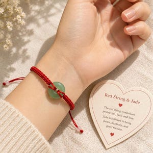 May include: A red string bracelet featuring a smooth, circular green jade stone at the center. The bracelet includes small beads at the ends of the adjustable cord. A heart-shaped card is present with the text Red String & Jade and descriptive text.