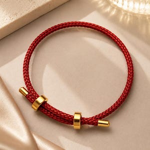 May include: A red braided bracelet with gold-colored metal accents. The bracelet features a circular design with two gold rings and two gold-colored end pieces. The jewelry is displayed on a neutral-toned surface.