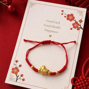 May include: A red cord bracelet featuring a central gold-colored charm shaped like a stylized animal, flanked by two small gold-colored beads. The bracelet is displayed on a white card printed with red floral illustrations and the text Good Luck Good Health Happiness.