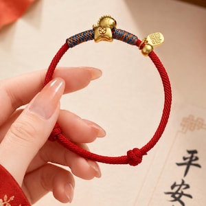 May include: A red cord bracelet featuring a gold-toned charm shaped like a stylized animal, accented with blue and red woven thread details and a small gold-toned rectangular tag with engraved characters, finished with two adjustable sliding knots.