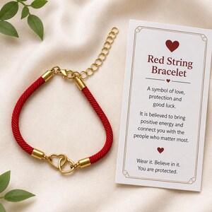 May include: A red string bracelet with gold-colored accents and a heart-shaped clasp. The bracelet is displayed with a card that reads "Red String Bracelet" and describes its symbolism of love, protection, and good luck.