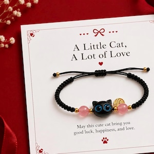 May include: A black cord bracelet featuring a central black cat bead with blue eyes, flanked by two translucent pink beads and small gold-tone accents. The display card reads: A Little Cat, A Lot of Love. May this cute cat bring you good luck, happiness, and love.