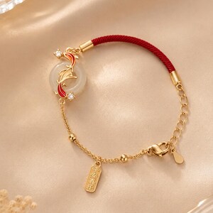 May include: Gold-toned bracelet featuring a red cord and a decorative pendant. The pendant includes a white circular stone with a gold and red fish design, and a small rectangular charm with Chinese characters.