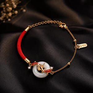 May include: A gold-tone bracelet featuring a white circular stone charm accented with red and gold fish-shaped metalwork and two small clear stones. The band consists of a red cord section and a gold-tone chain with a small rectangular metal tag.