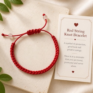 May include: A red string knot bracelet with a braided design. The bracelet has a sliding knot closure and two small, round, clear beads. A card next to the bracelet reads "Red String Knot Bracelet".