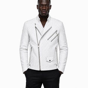 May include: A white, textured leather jacket with a snake-skin pattern. The jacket features a diagonal zipper, multiple silver zippers, and a small pocket. The model is wearing black pants. The jacket has a classic biker style.