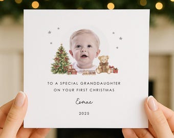 May include: A square white greeting card featuring a central photograph of a baby surrounded by illustrations of a Christmas tree, a teddy bear, and gifts. The text reads: TO A SPECIAL GRANDDAUGHTER ON YOUR FIRST CHRISTMAS, Esmae, 2025.