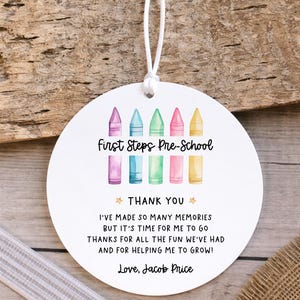 May include: A round white ornament with a white string featuring a watercolour illustration of five colourful crayons and the text First Steps Pre-School, THANK YOU, I'VE MADE SO MANY MEMORIES BUT IT'S TIME FOR ME TO GO THANKS FOR ALL THE FUN WE'VE HAD AND FOR HELPING ME TO GROW! Love, Jacob Price.