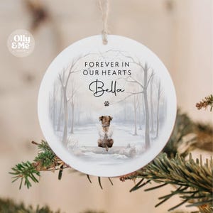 May include: A white, circular Christmas decoration with the words "FOREVER IN OUR HEARTS" and the name "Bella". It shows a watercolour illustration of a dog in a snowy forest. A paw print is below the name.