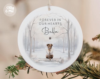 May include: A white, round Christmas ornament with the text "FOREVER IN OUR HEARTS" and the name "Bella". It features a watercolor illustration of a dog in a snowy forest scene. A paw print is below the name.