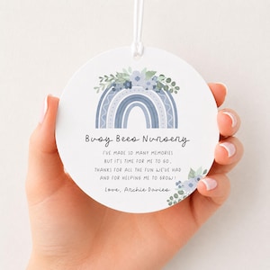 May include: A round white ornament featuring a blue rainbow graphic topped with blue flowers and green leaves. The text reads: Busy Bees Nursery, I've made so many memories but it's time for me to go, thanks for all the fun we've had and for helping me to grow! Love, Archie Davies.