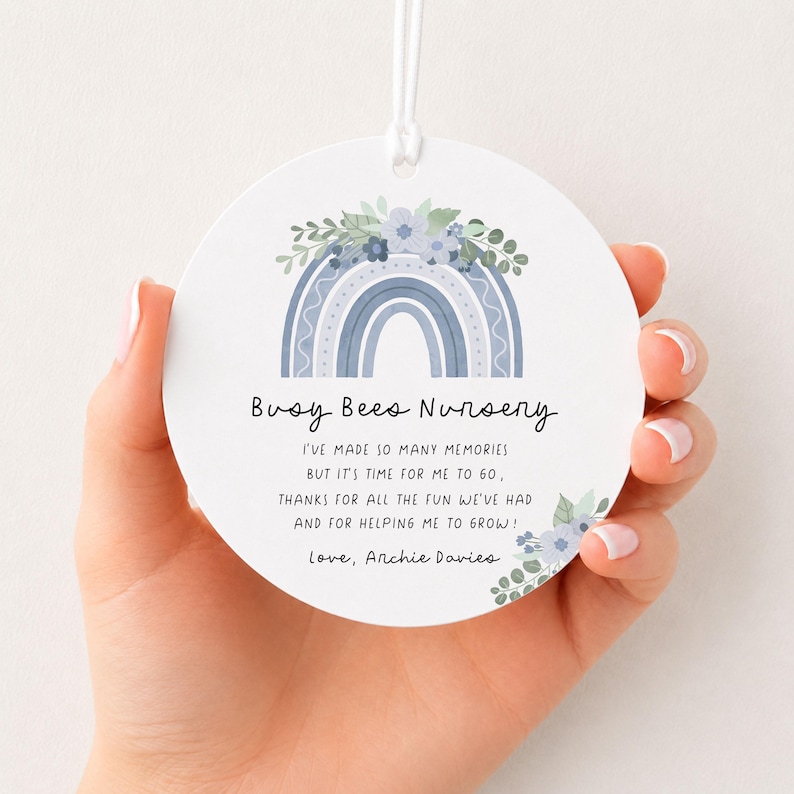 May include: A round white ornament featuring a blue rainbow graphic topped with blue flowers and green leaves. The text reads: Busy Bees Nursery, I've made so many memories but it's time for me to go, thanks for all the fun we've had and for helping me to grow! Love, Archie Davies.