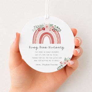 May include: A round white ceramic ornament featuring a watercolor rainbow graphic topped with pink and green flowers. The text reads: Busy Bees Nursery, I've made so many memories but it's time for me to go, thanks for all the fun we've had and for helping me to grow! Love, Archie Davies.