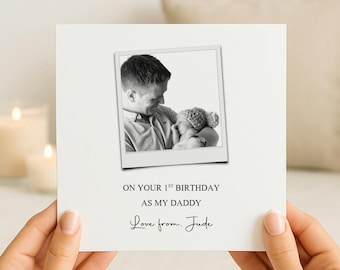 May include: A square white greeting card featuring a black and white photograph of a parent holding a baby. The text reads: ON YOUR 1ST BIRTHDAY AS MY DADDY. Below this, the text reads: Love from. Jude. The card has a clean, minimalist design.