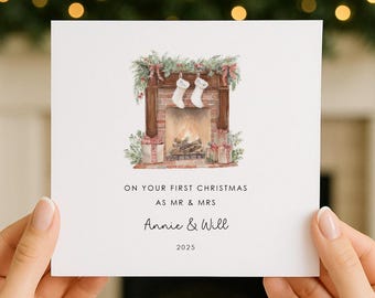 May include: A square greeting card featuring a watercolor illustration of a brick fireplace with two white stockings, greenery, and wrapped gifts. The text reads: ON YOUR FIRST CHRISTMAS AS MR & MRS, Annie & Will, 2025, in black lettering.