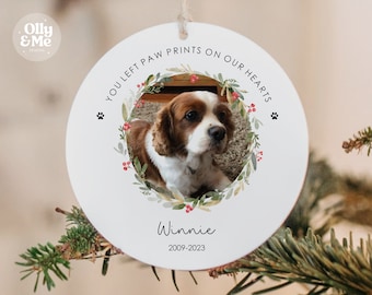 May include: White round Christmas ornament with a photo of a brown and white dog inside a green and red wreath. The ornament says "You left paw prints on our hearts" and "Winnie 2009-2023".