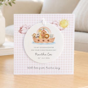 May include: A square greeting card with a light pink gingham pattern featuring a circular white ornament hanging from a white string. The ornament displays a watercolour illustration of a wooden ark with animals, a rainbow, and the text: TO MY GODDAUGHTER ON YOUR CHRISTENING DAY, Martha Eve, 26TH JULY 2026. The card bottom reads: With love from, Auntie Lucy.