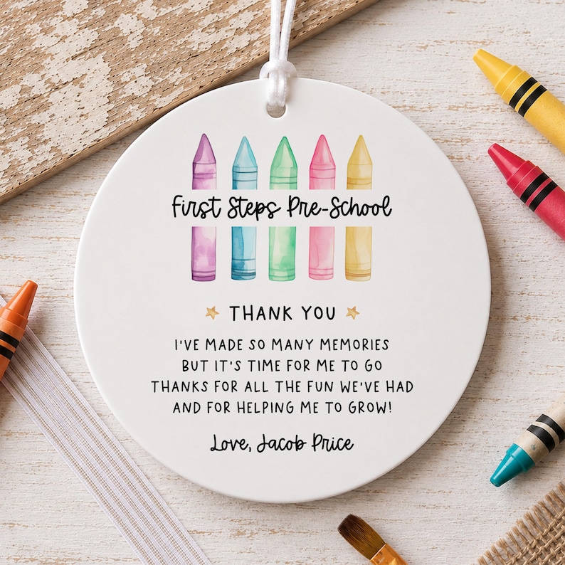 May include: A round white ceramic ornament with a white hanging string. The design features five watercolour-style crayons in purple, blue, green, pink, and yellow above the text First Steps Pre-School, Thank You, and a poem ending with Love, Jacob Price.