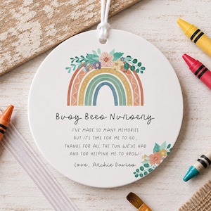 May include: A round white ceramic ornament featuring a colorful rainbow illustration topped with flowers. The text reads: Busy Bees Nursery, I've made so many memories but it's time for me to go, thanks for all the fun we've had and for helping me to grow! Love, Archie Davies.